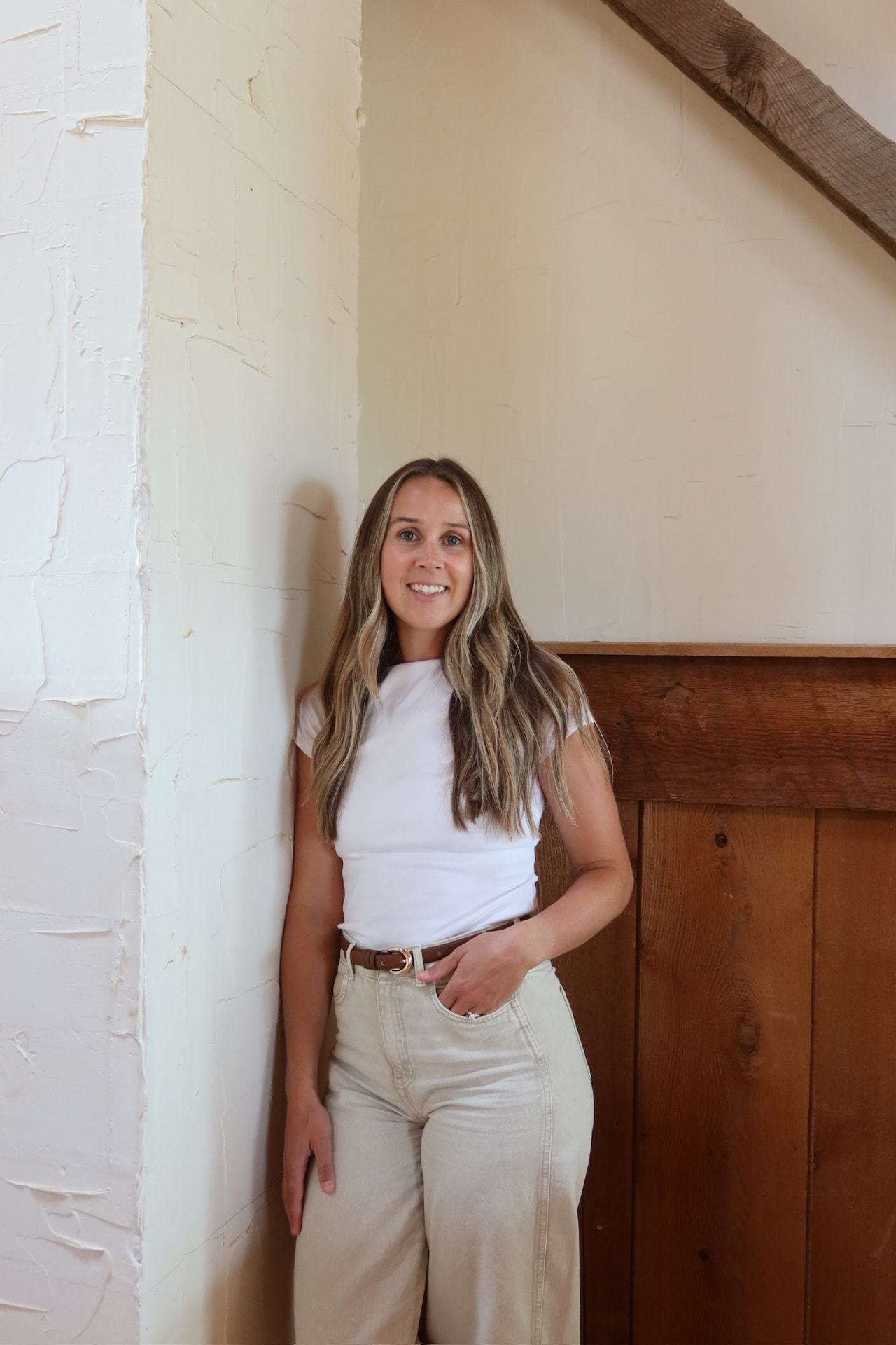 Aly, founder of Tide Together Consulting, standing in a bright minimal space with textured walls and a wooden beam