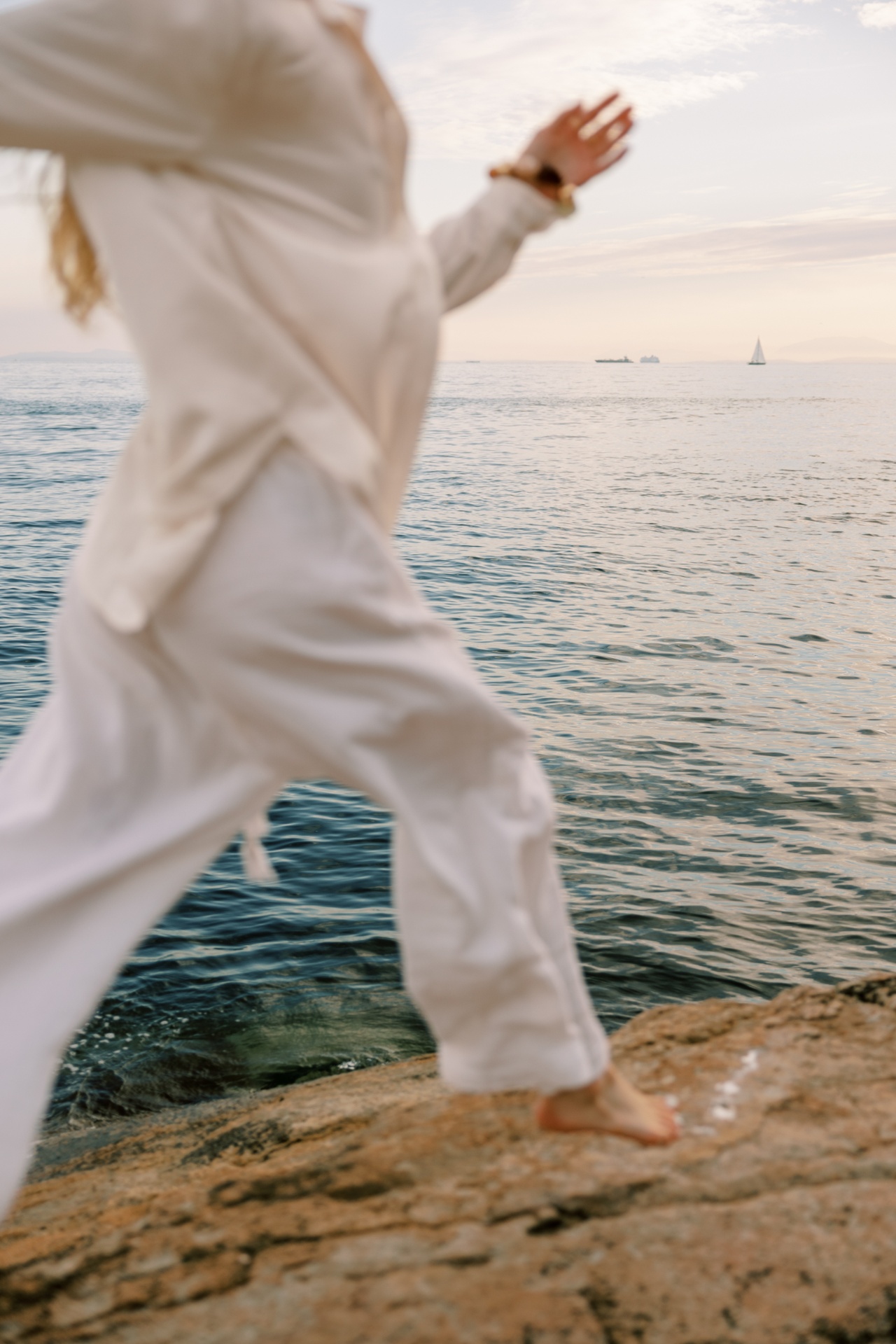 A woman in flowing white linen walking along a rocky shoreline at golden hour